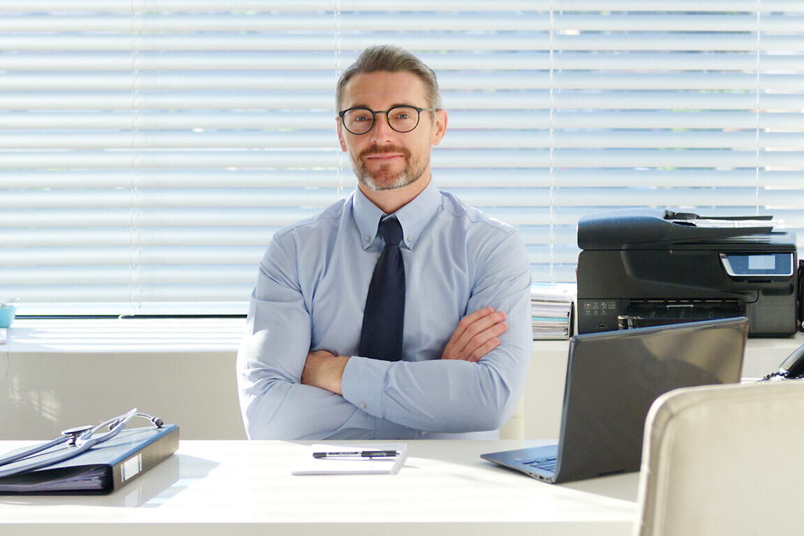 Portrait Of Smiling Mature Male Doctor Sitting Behind Desk In Office Or Surgery