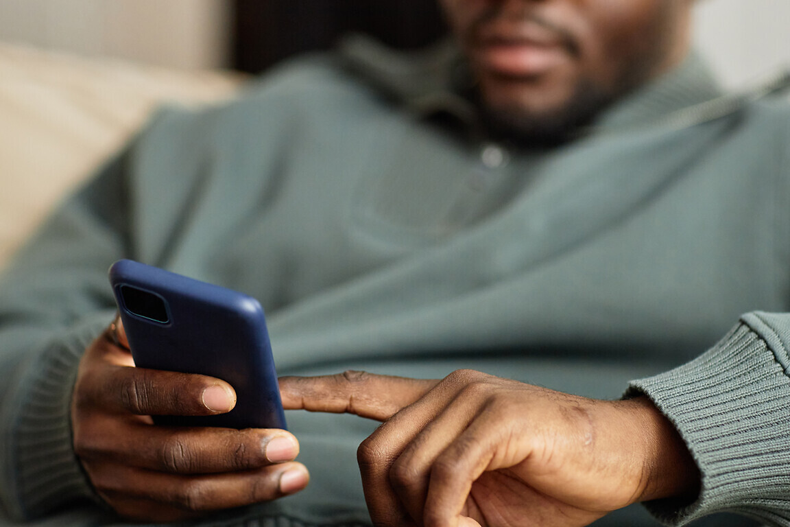 Close up of man holding phone in hand and tapping screen with finger while relaxing on couch at home, copy space