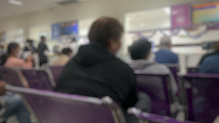 Blurred image of a large number of patients sitting in a large hospital waiting room.