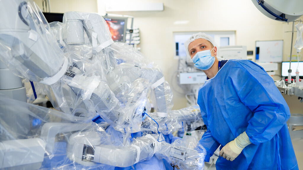 Surgeon using robot arm in OR. A surgeon in blue scrubs uses a robotic arm during a surgical procedure in a modern operating room.