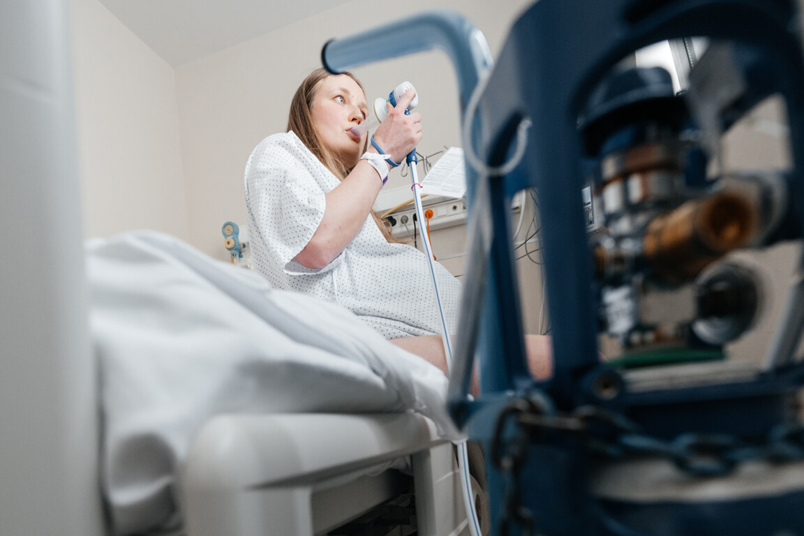 A pregnant woman in a hospital gown sits on a bed, inhaling gas and air for pain relief during labour.