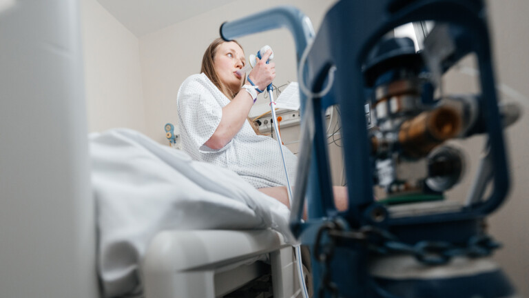 A pregnant woman in a hospital gown sits on a bed, inhaling gas and air for pain relief during labour.