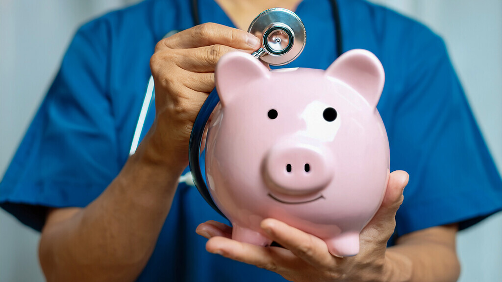 Nurse or doctor with stethoscope holding piggy bank in hand.