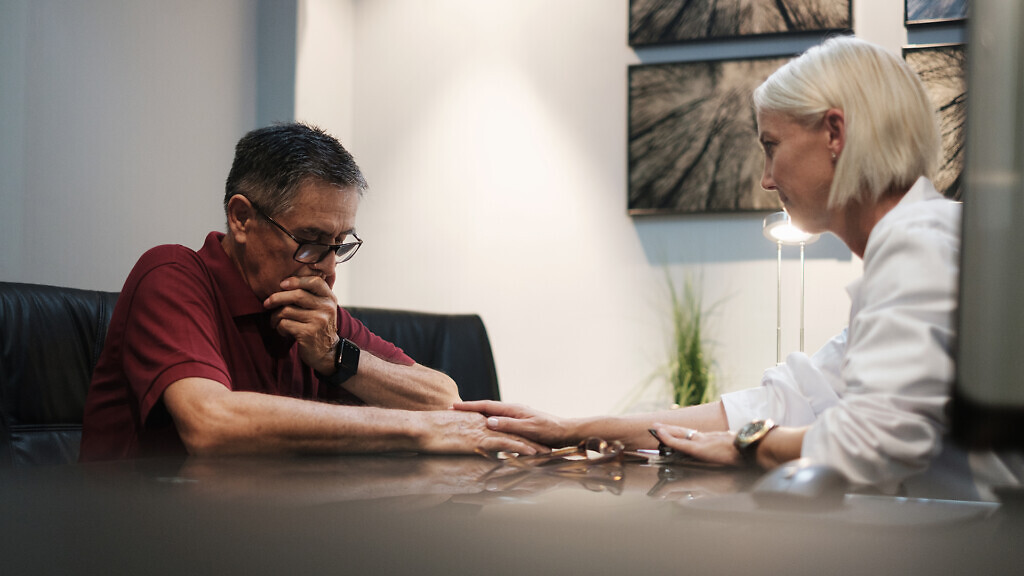 An emotional worried senior man with serious illness gets a bad diagnosis from compassionate doctor in hospital. Caucasian female health specialist showing emphaty towards patient while touching hand