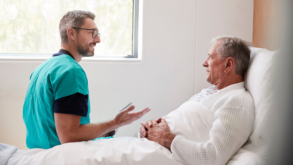 Surgeon Visiting And Talking With Senior Male Patient In Hospital Bed In Geriatric Unit