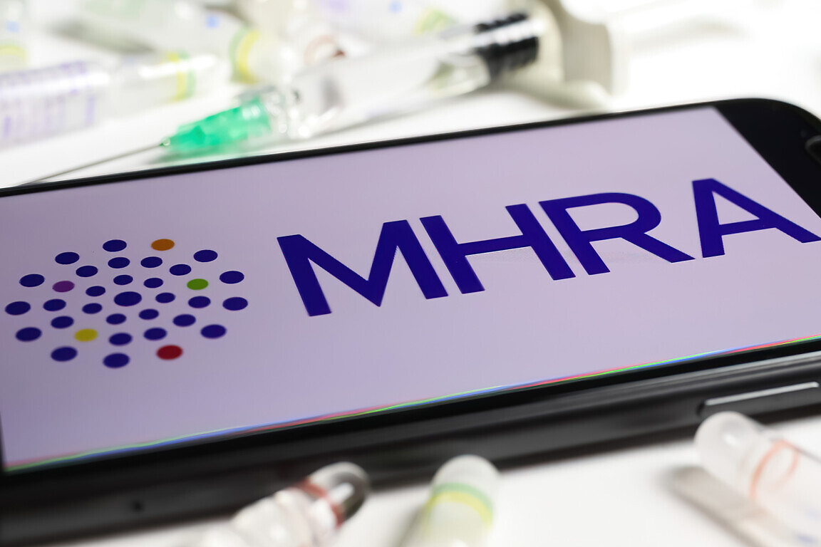 Closeup of mobile phone screen with logo lettering of UK MHRA agency, serum vials and syringe background