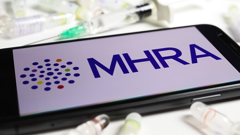 Closeup of mobile phone screen with logo lettering of UK MHRA agency, serum vials and syringe background