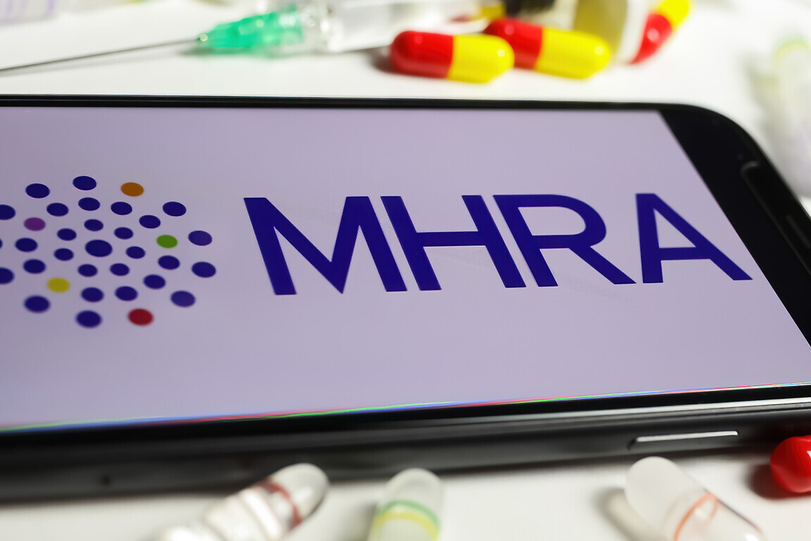 Closeup of mobile phone screen with logo lettering of UK MHRA agency, serum vials and syringe background