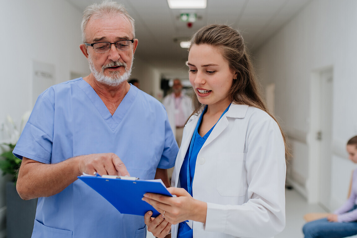 Older doctor giving advise to his younger colleague, discussing at a hospital corridor. Health care concept.