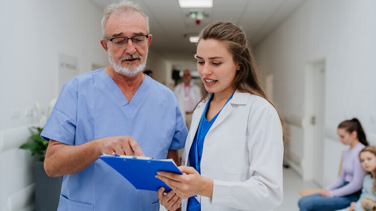 Older doctor giving advise to his younger colleague, discussing at a hospital corridor. Health care concept.