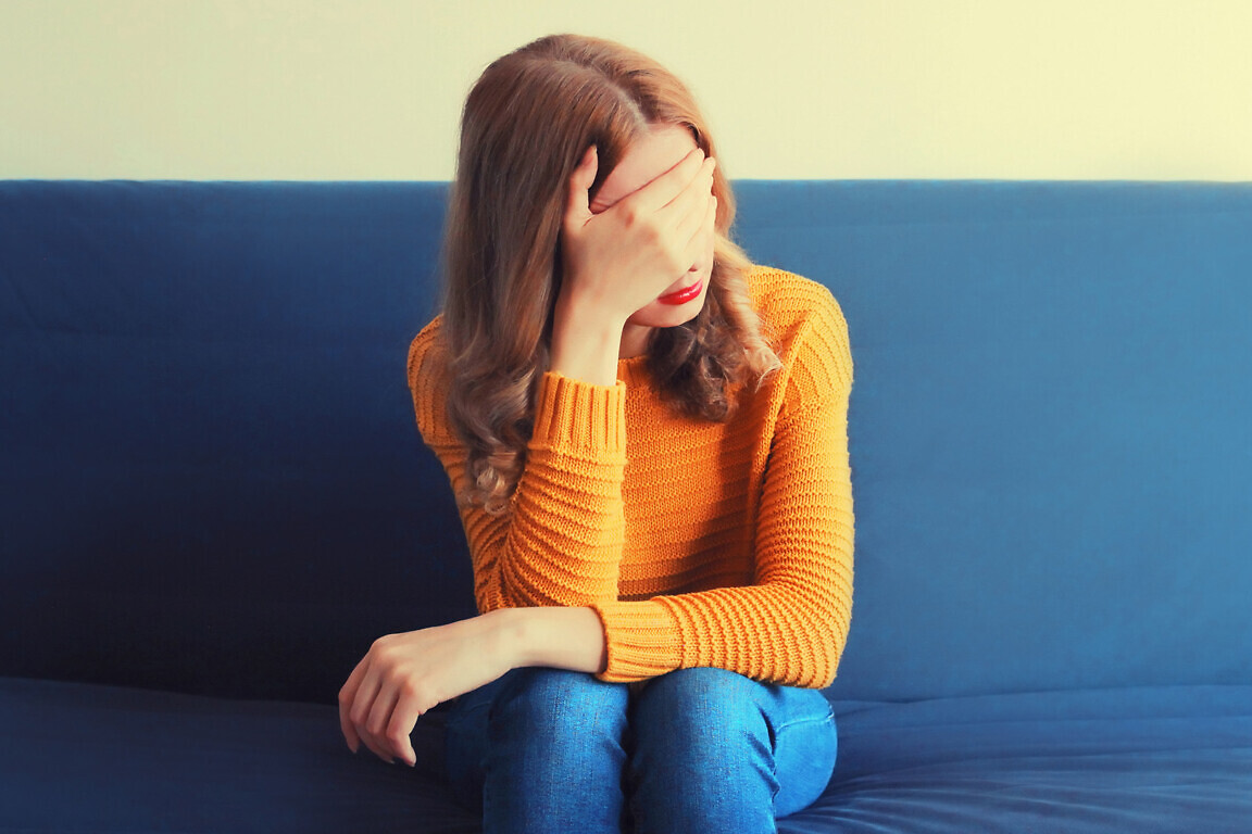 Upset sad crying woman covering her eyes with her hands while experiencing mental or physical suffering on the couch at home