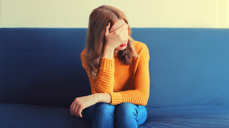 Upset sad crying woman covering her eyes with her hands while experiencing mental or physical suffering on the couch at home
