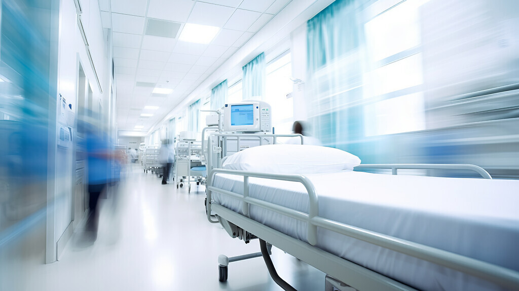 A hospital bed in the corridor of a busy hospital with people walking past.