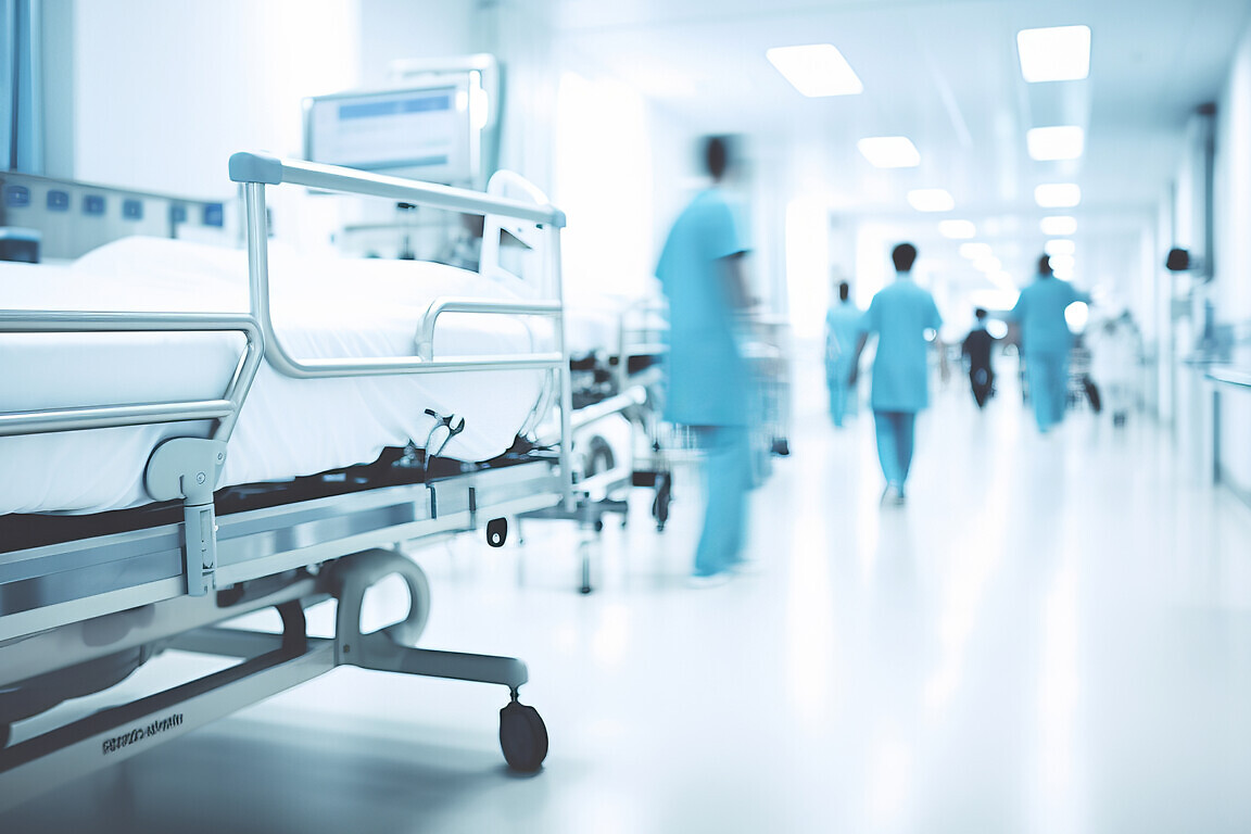 A hospital bed in the corridor of a busy hospital with people walking past.