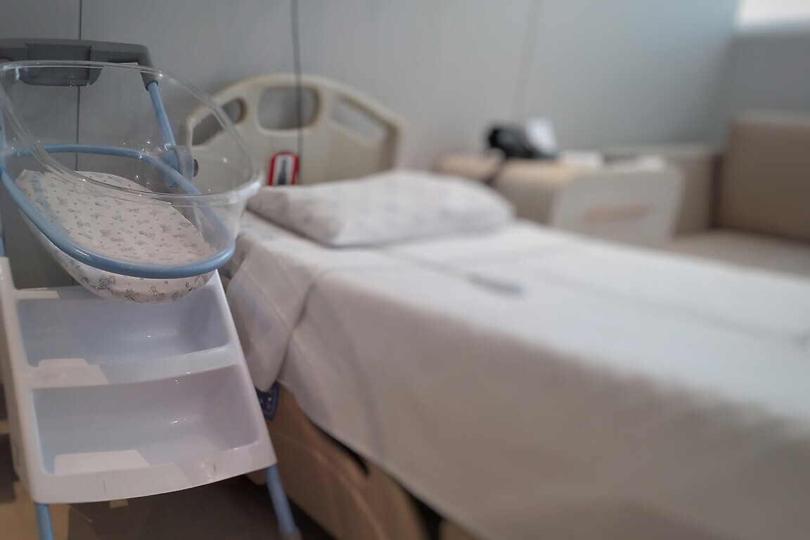 Acrylic crib for newborn, next to the bed in a maternity room.