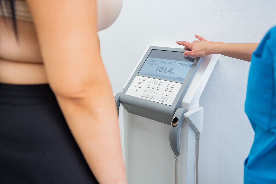 Nurse aids a overweight female patient in measuring weight on a digital scale during a healthcare appointment