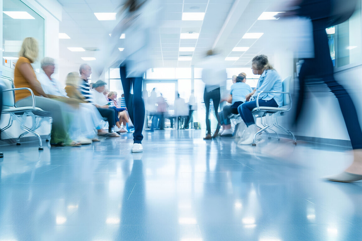 A busy hospital waiting area with patients sitting in chairs and blurred medical staff walking through the corridor, capturing the hustle of a healthcare environment