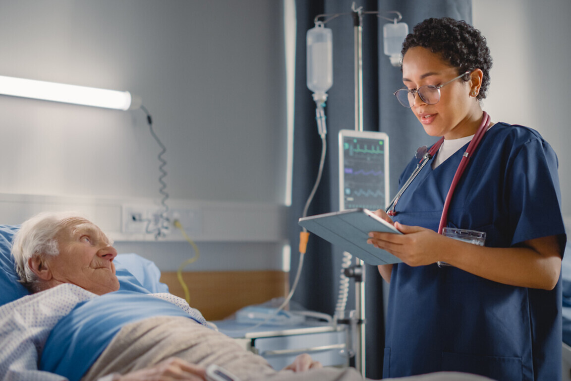Hospital Ward: Friendly Head Nurse Talks with Elderly Patient Resting in Bed. Physician Uses Tablet Computer, Does Checkup, Ask Health Care Questions. Old Man Fully Recovering after Successful Surgery