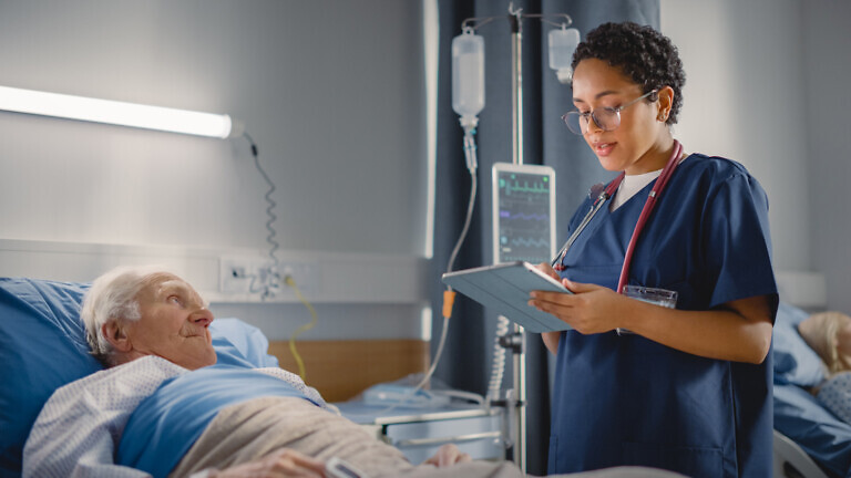 Hospital Ward: Friendly Head Nurse Talks with Elderly Patient Resting in Bed. Physician Uses Tablet Computer, Does Checkup, Ask Health Care Questions. Old Man Fully Recovering after Successful Surgery